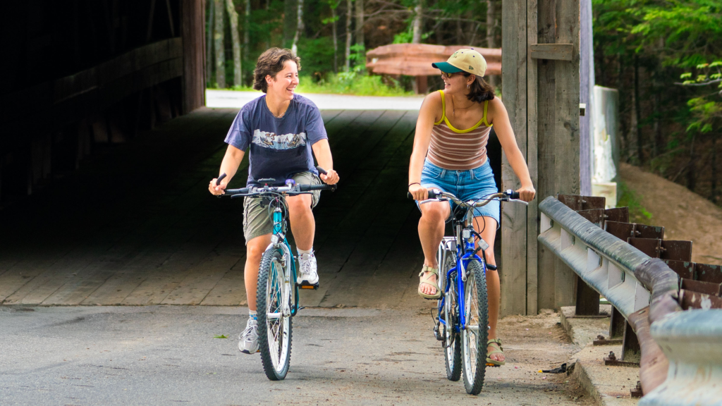 Two young women laughing while riding bicycles through a covered bridge in Portland Maine.