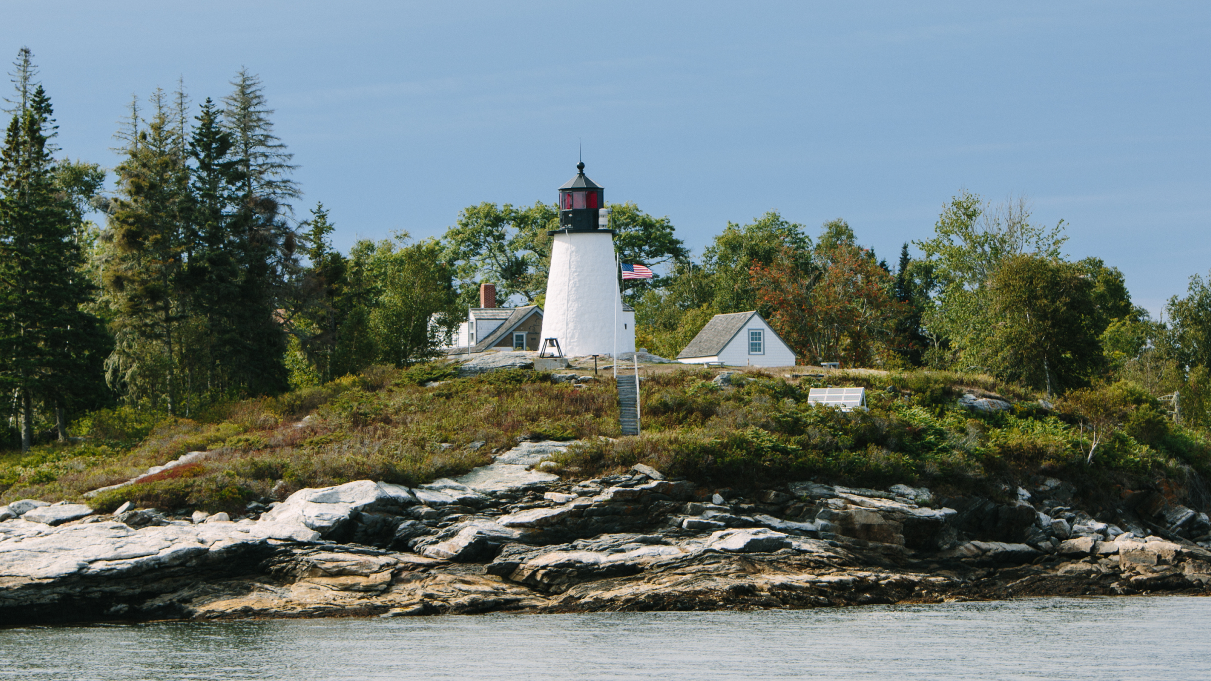 A white lighthouse stands on a rocky island surrounded by trees and water in Portland Maine.