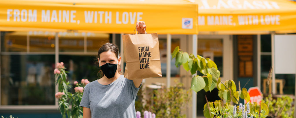 Woman holding up bag saying 'from Maine with love' while wearing mask, Photo Courtesy of Allagash Brewing and their staff