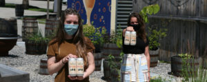 Two women in masks carrying cans of beer, Photo Courtesy of Mat Trogner, via Allagash Brewing