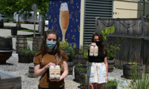 Two women in masks carrying cans of beer, Photo Courtesy of Mat Trogner, via Allagash Brewing
