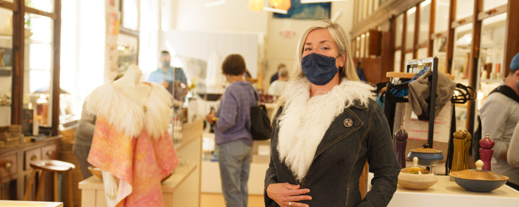 Woman in mask trying on coat at Maine Craft Portland, Photo Courtesy of Good Maine Woman in mask trying on coat at Maine Craft Portland, Photo Courtesy of Good Maine