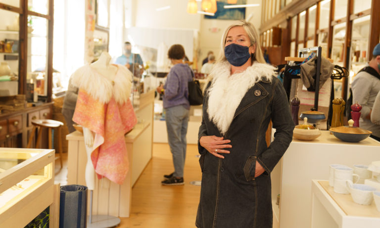 Woman in mask trying on coat at Maine Craft Portland, Photo Courtesy of Good Maine Woman in mask trying on coat at Maine Craft Portland, Photo Courtesy of Good Maine