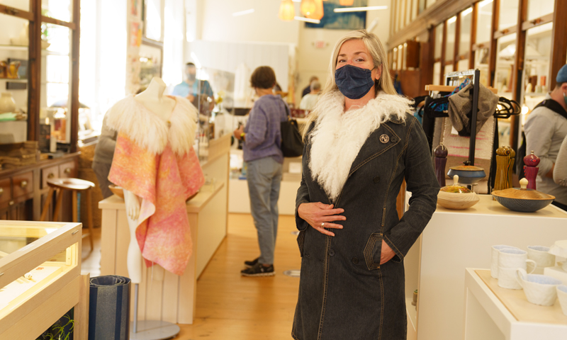 Woman in mask trying on coat at Maine Craft Portland, Photo Courtesy of Good Maine