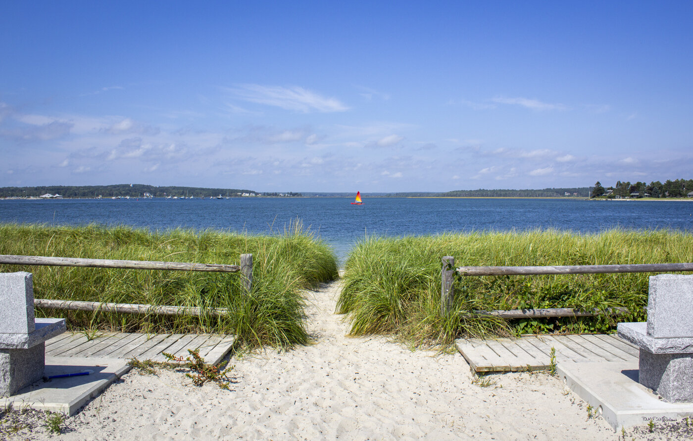 A sandy path leads through beach grass to a calm blue bay with a sailboat in Portland Maine.