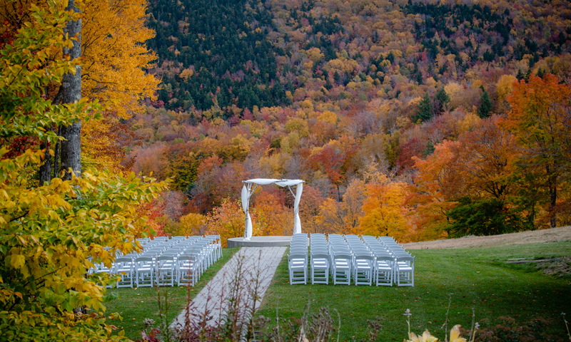 An outdoor wedding setup with chairs facing an arch against a vibrant autumn forest in Portland Maine.