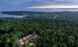 Aerial of yurt community surrounded by green woods and ocean at sunset; Photo Credit: Peter G. Morneau Photography