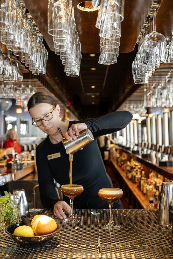 A bartender pours a cocktail into a glass at a bar with hanging glasses in Portland Maine.