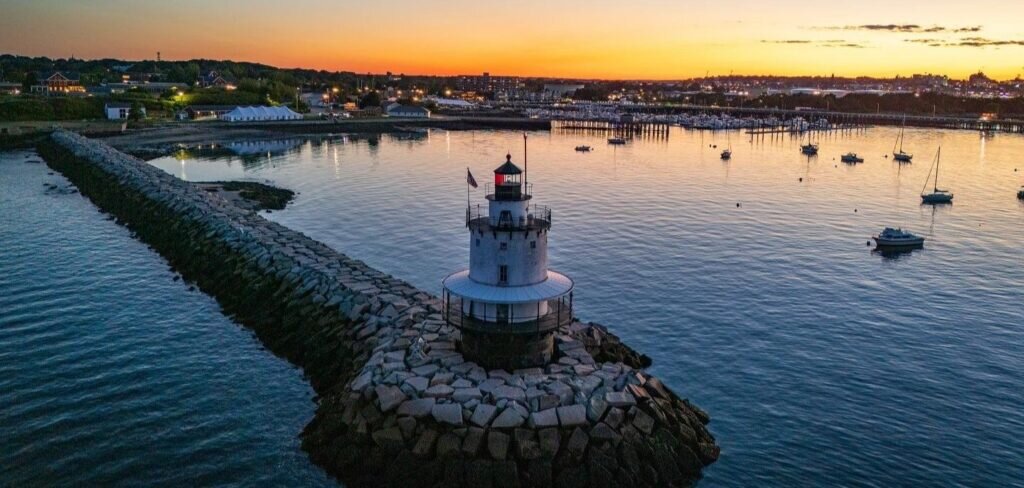 A lighthouse stands on a stone breakwater in a harbor at sunset in Portland Maine.