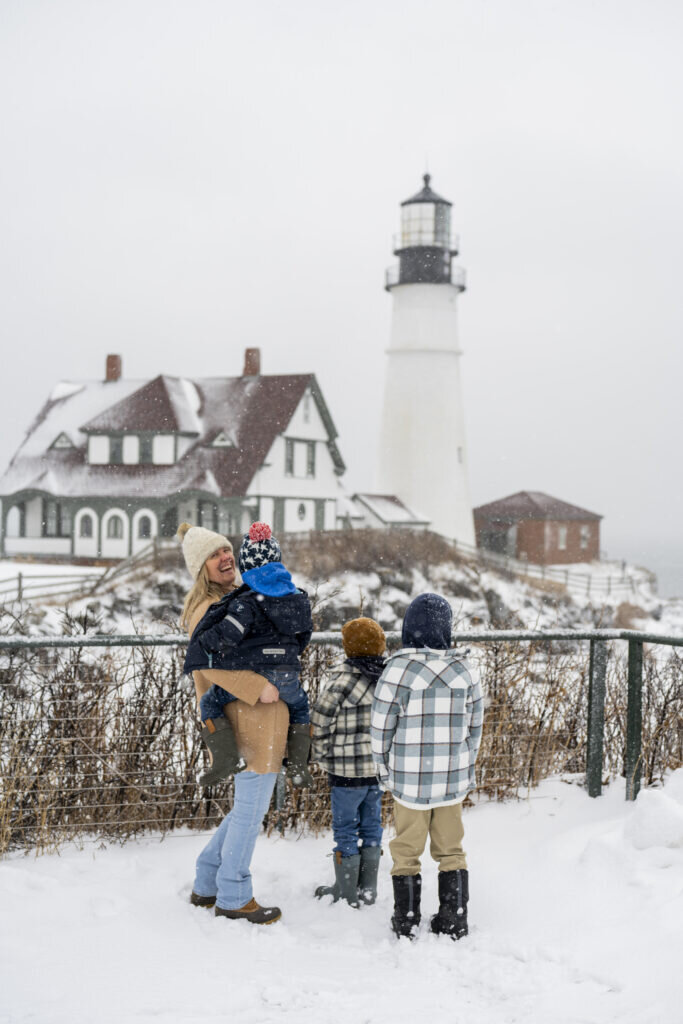 A woman and three children stand in the snow looking at a lighthouse in Portland Maine.