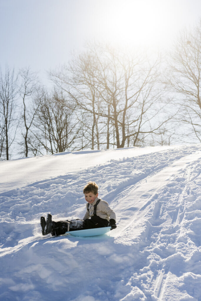 A young boy happily sledding down a snow-covered hill on a sunny day in Portland Maine.