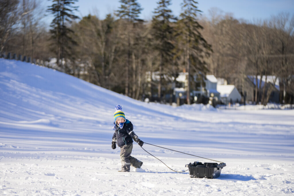 A young child in winter gear pulls a black sled across a snowy field in Portland Maine.