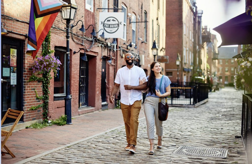 A diverse couple walks arm-in-arm down a historic cobblestone street in Portland Maine.