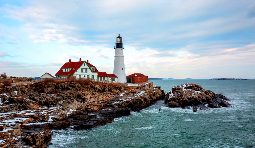 A white lighthouse stands on a rocky cliff overlooking the ocean under a cloudy sky in Portland Maine.