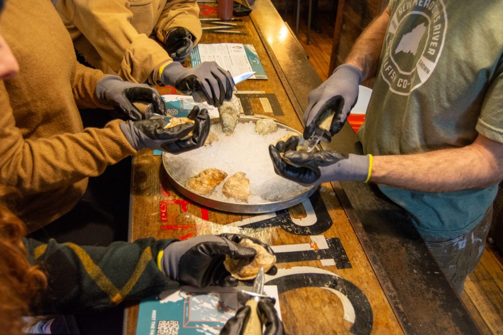 People wearing protective gloves shuck oysters on a bed of ice at a counter in Portland Maine.