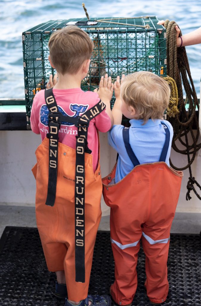 Two young children in orange bib overalls look at a lobster trap on a boat in Portland Maine.