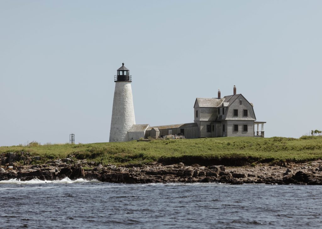 A white lighthouse and an attached house on a grassy coastal island in Portland Maine.