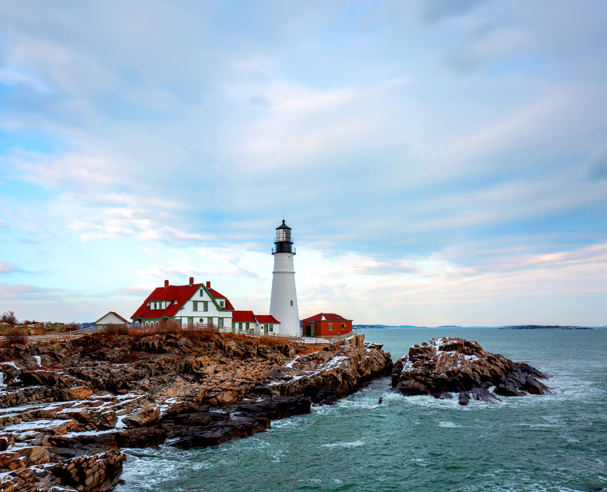 A white lighthouse stands on a rocky coast with a house nearby in Portland Maine.
