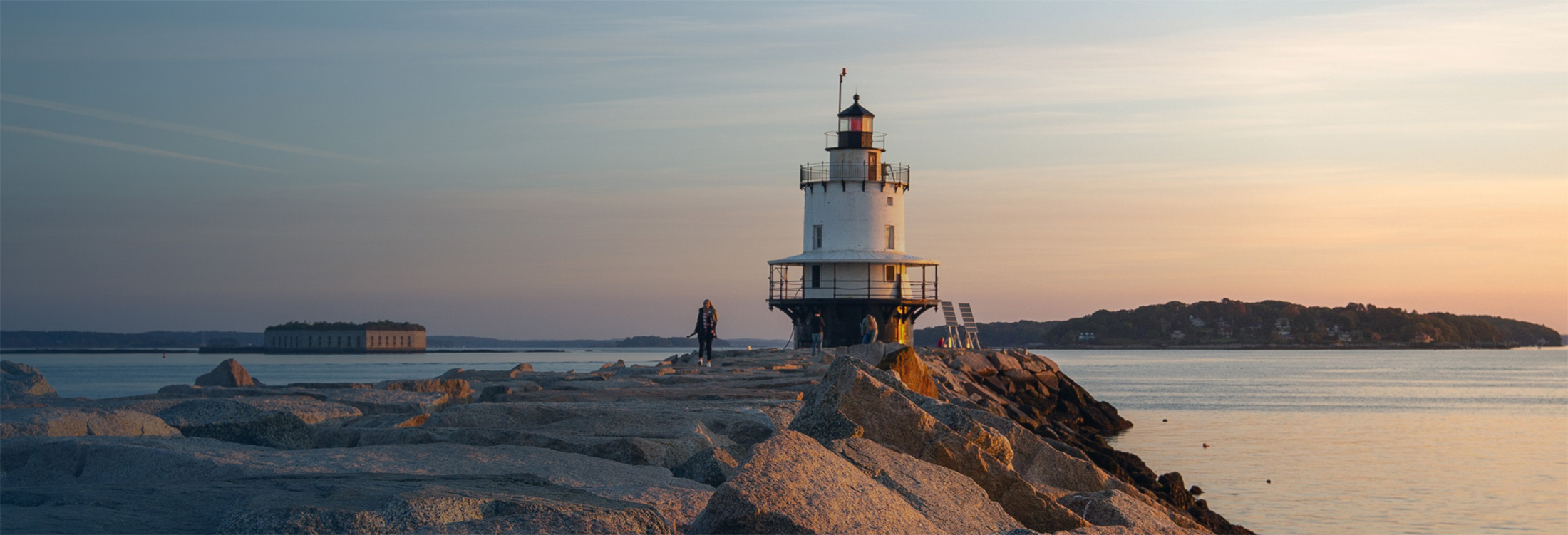 A white lighthouse stands on a rocky breakwater at sunset in Portland Maine.