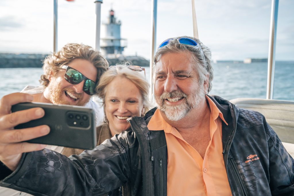 Three people smiling while taking a selfie on a boat with a lighthouse behind in Portland Maine.
