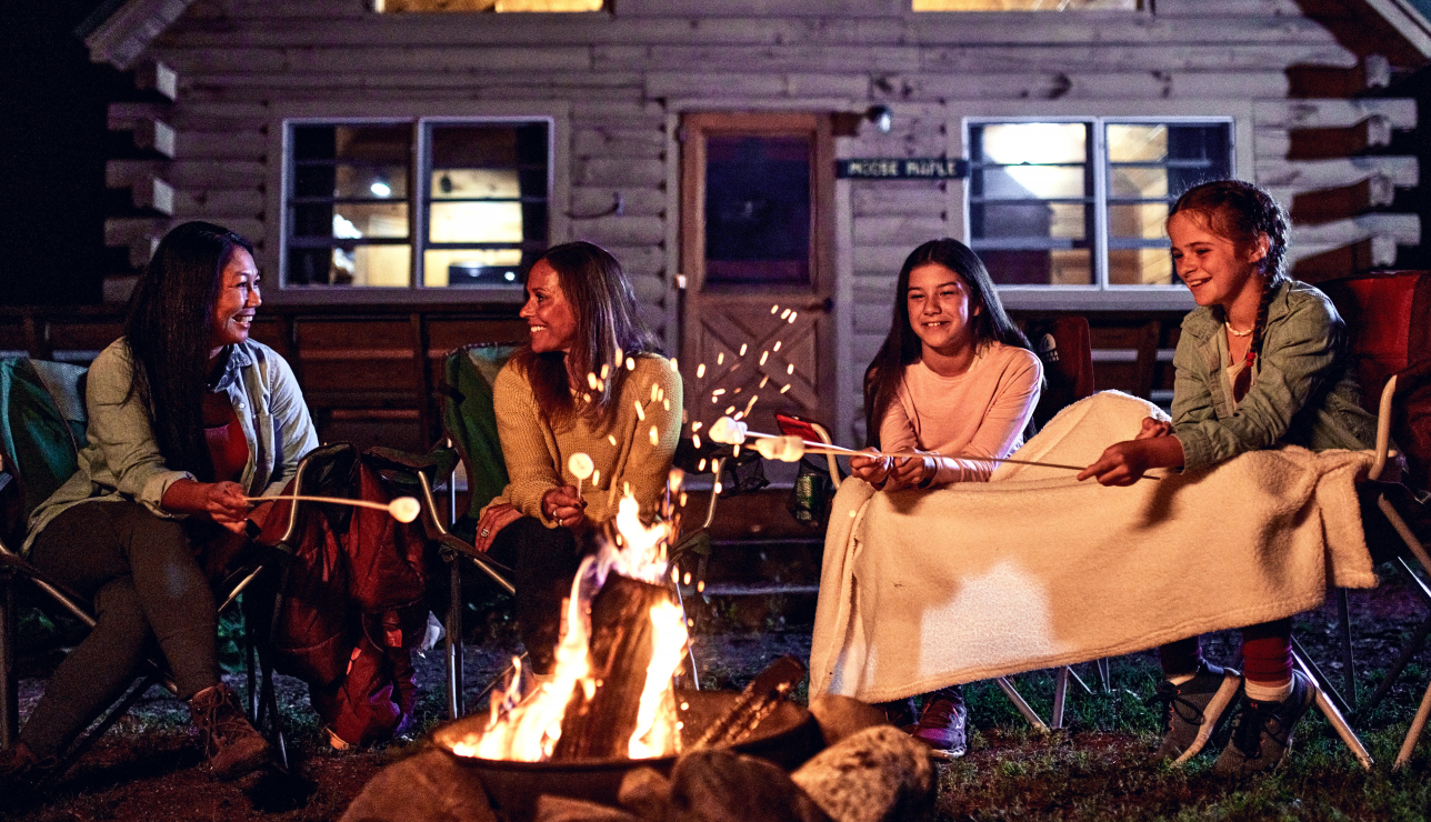 Four people sitting around a campfire roasting marshmallows in front of a log cabin in Portland Maine.