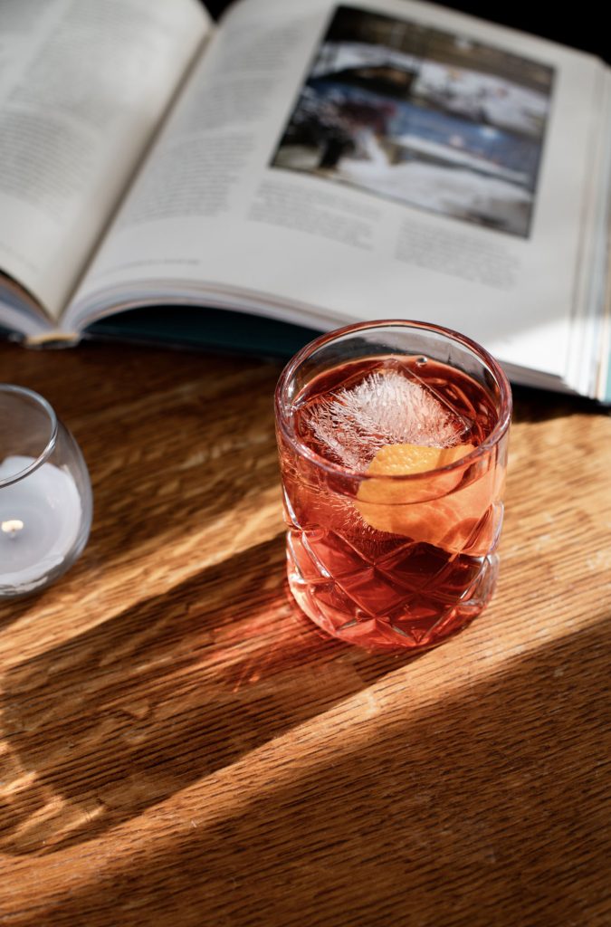 A red cocktail with ice and orange peel sits on a wooden table near books in Portland Maine.