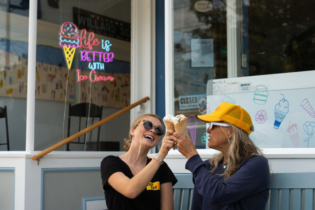 Two women laughing and clinking ice cream cones together in front of an ice cream shop in Portland Maine.