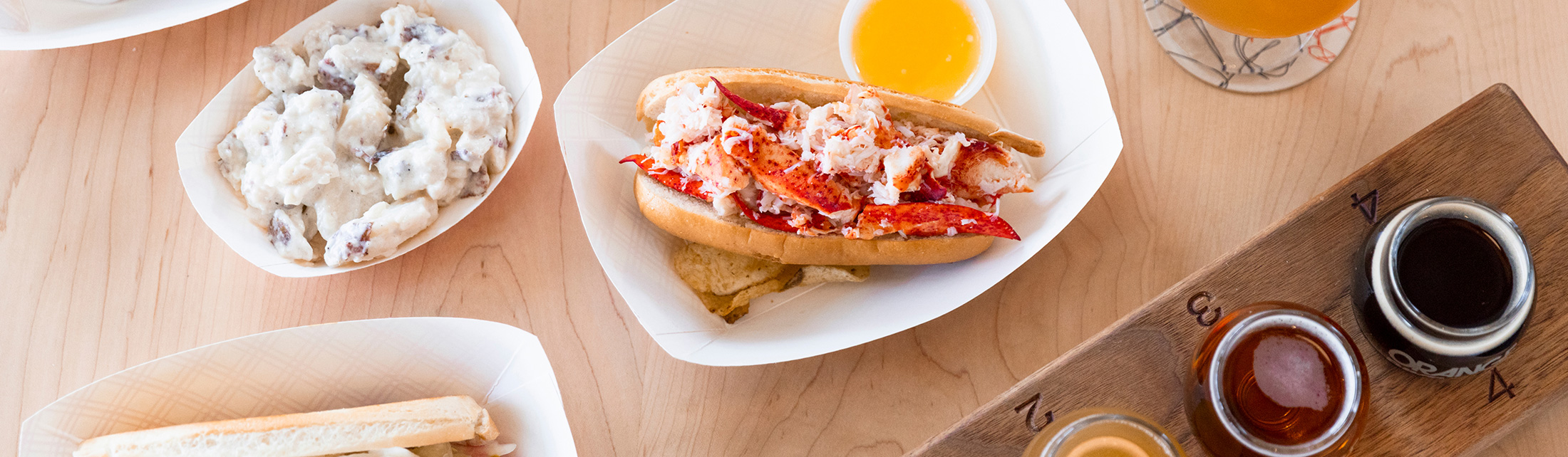 A lobster roll, potato salad, and a beer flight on a wooden table in Portland Maine.