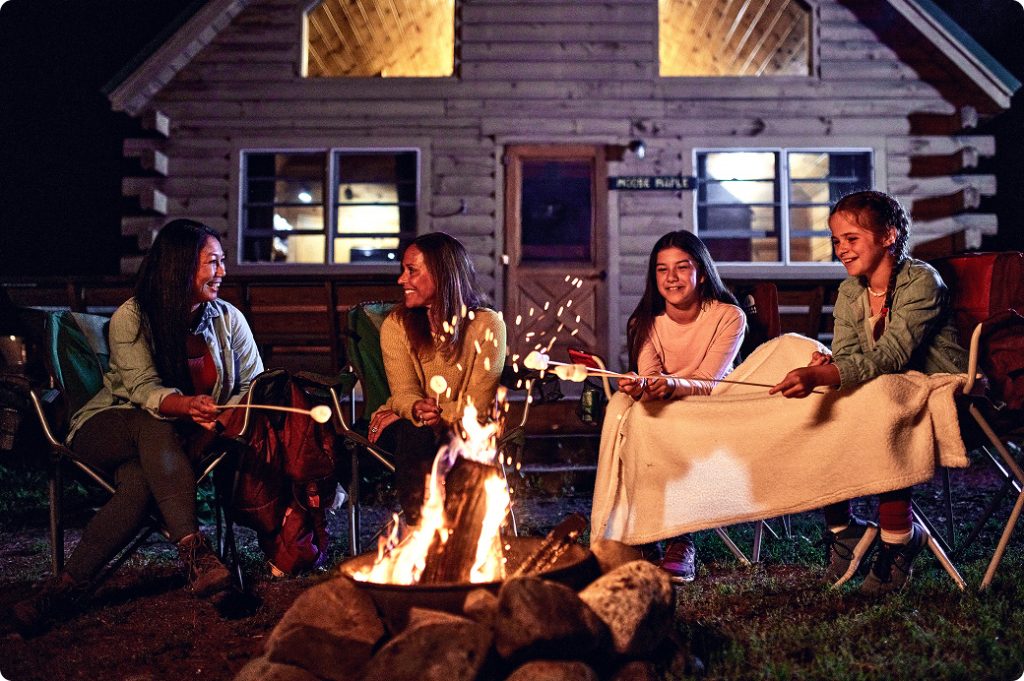 Four people sitting around a campfire roasting marshmallows in front of a log cabin in Portland Maine.