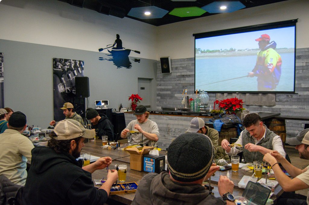 A group of men sitting at tables in a room, tying fishing flies together in Portland Maine.
