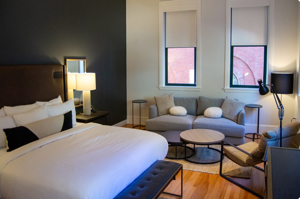 A modern hotel room featuring a bed, gray sofa, and two windows in Portland Maine.