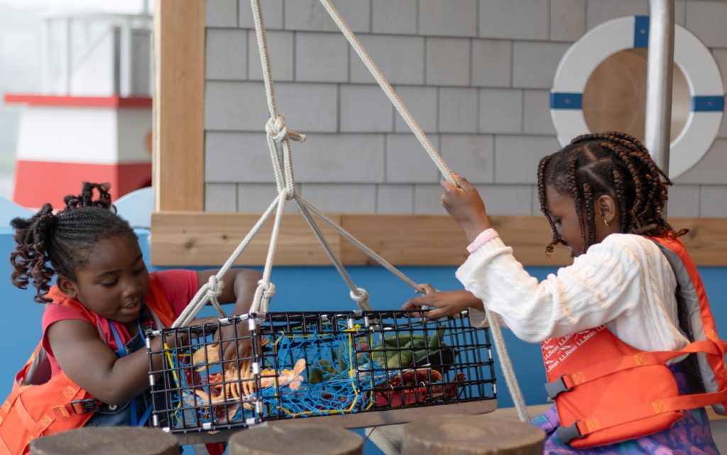 Two young girls playing with a toy lobster trap in a nautical-themed play area in Portland Maine.