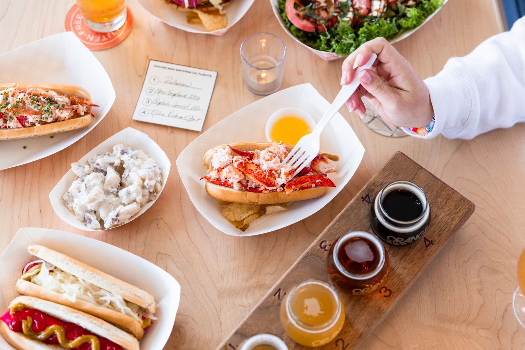 A person eating a lobster roll at a table with beer flights and sides in Portland Maine.