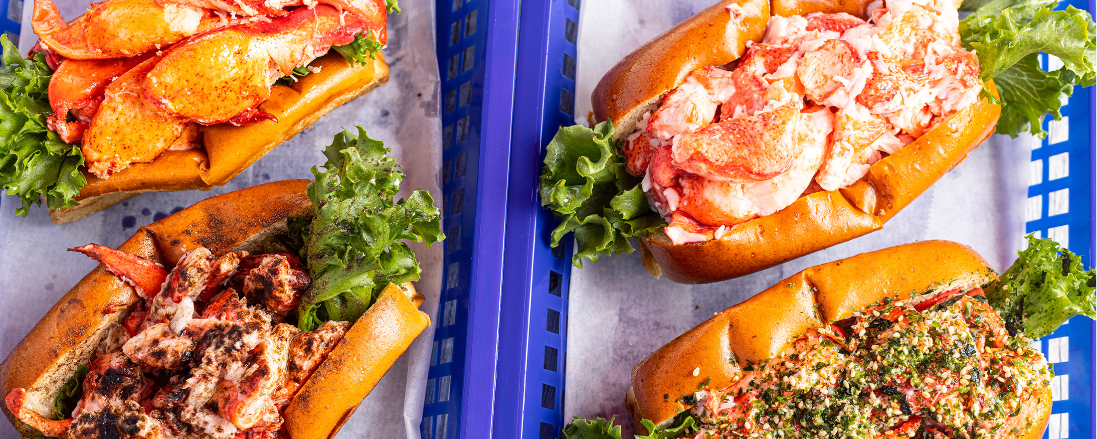Four lobster rolls served in blue plastic baskets on parchment paper in Portland Maine.