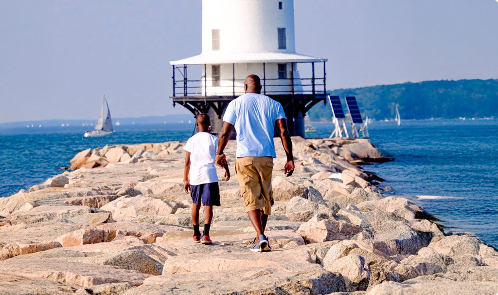 A father and son walk along a rocky breakwater toward a white lighthouse in Portland Maine.