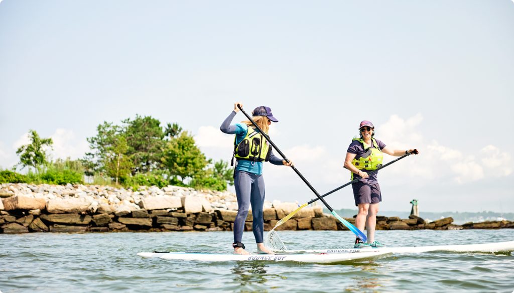 Two women paddleboarding on calm water near a rocky shoreline in Portland Maine.