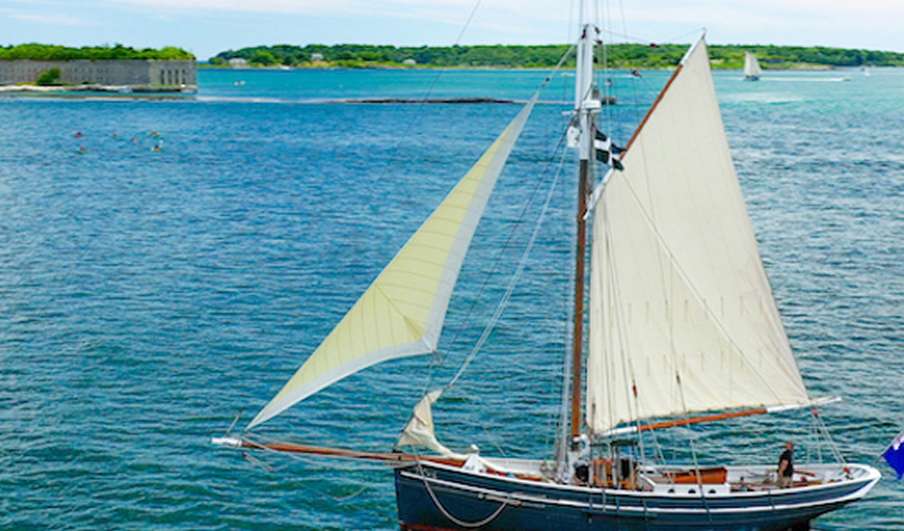 A sailboat with white sails cruises on blue water near a coastal fort in Portland Maine.