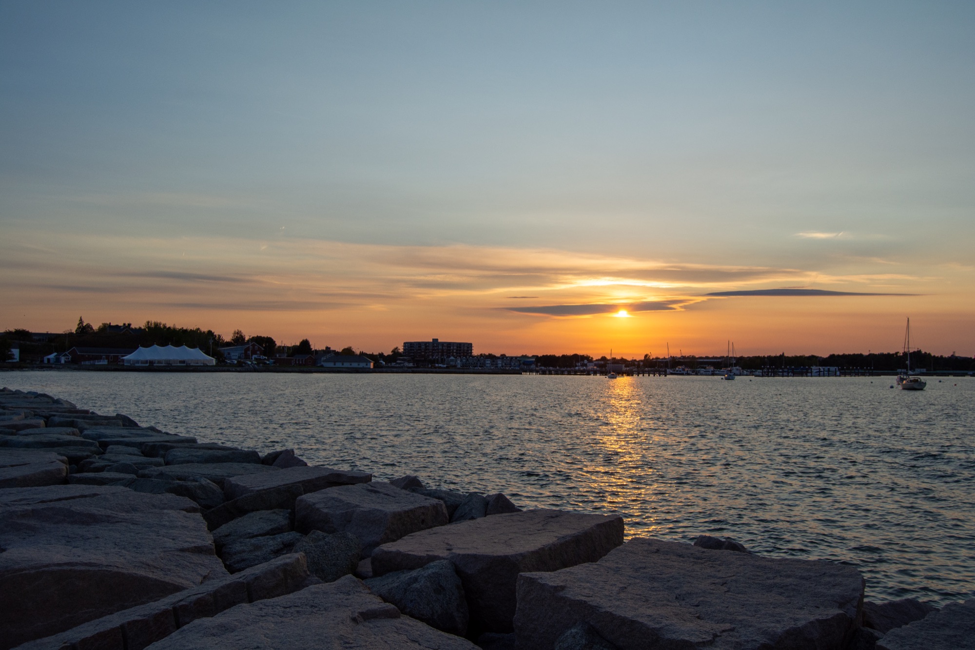 A calm harbor at sunset with rocky foreground and distant buildings in Portland Maine.