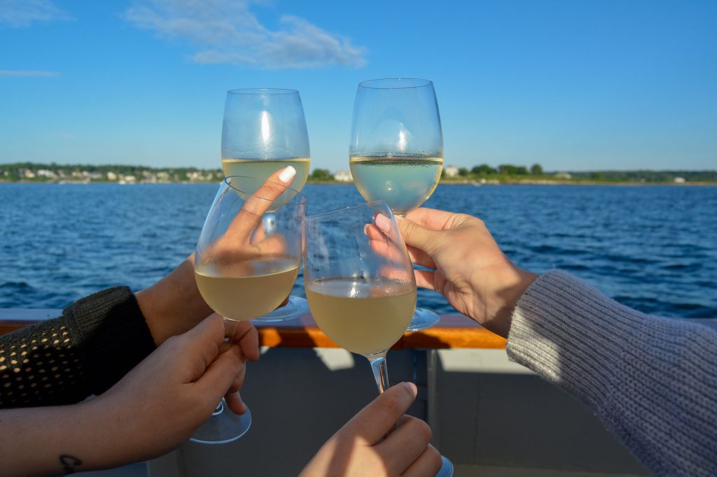 Four hands holding glasses of white wine for a toast on a boat in Portland Maine.