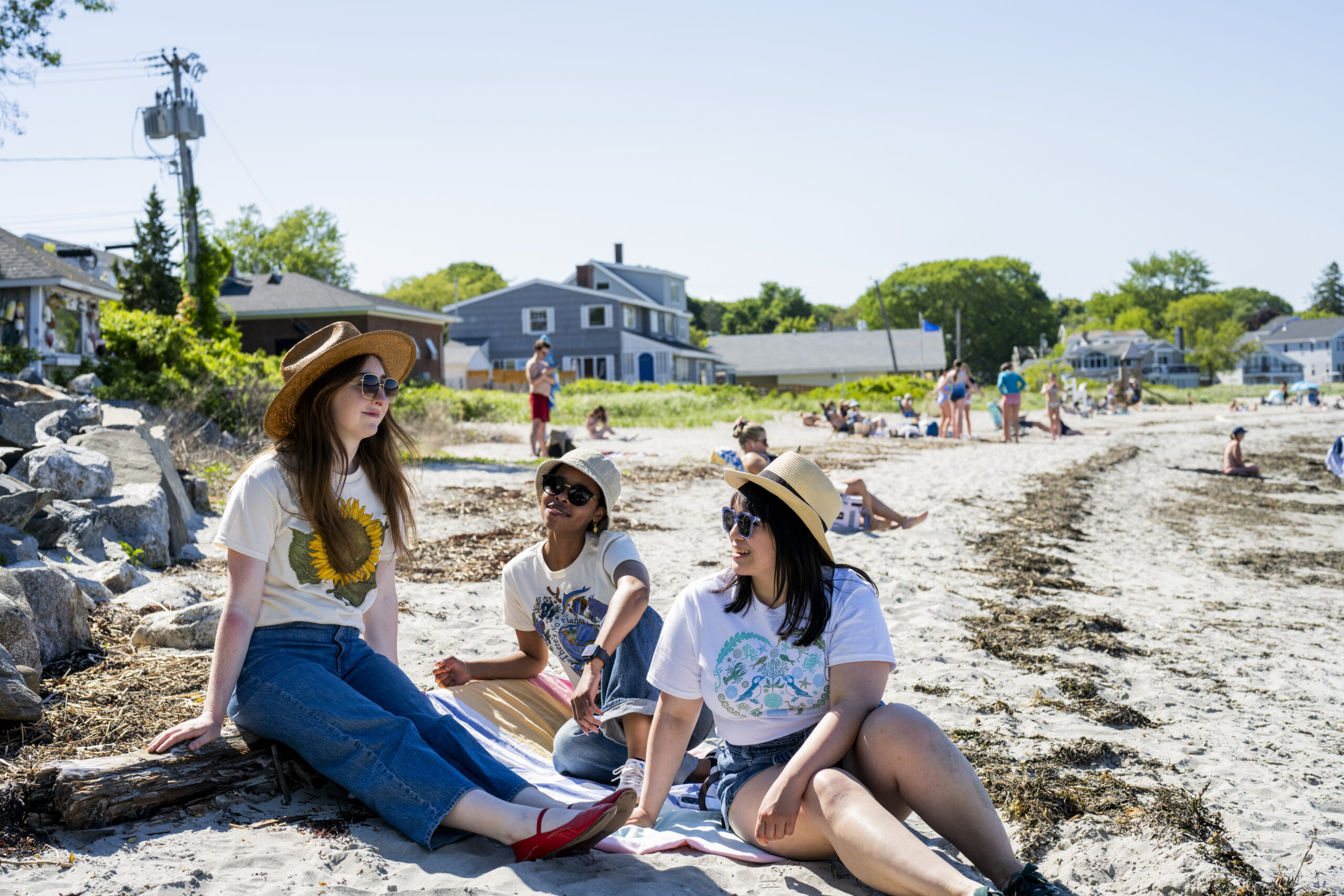 Three young women sitting on a sandy beach wearing hats and sunglasses in Portland Maine.