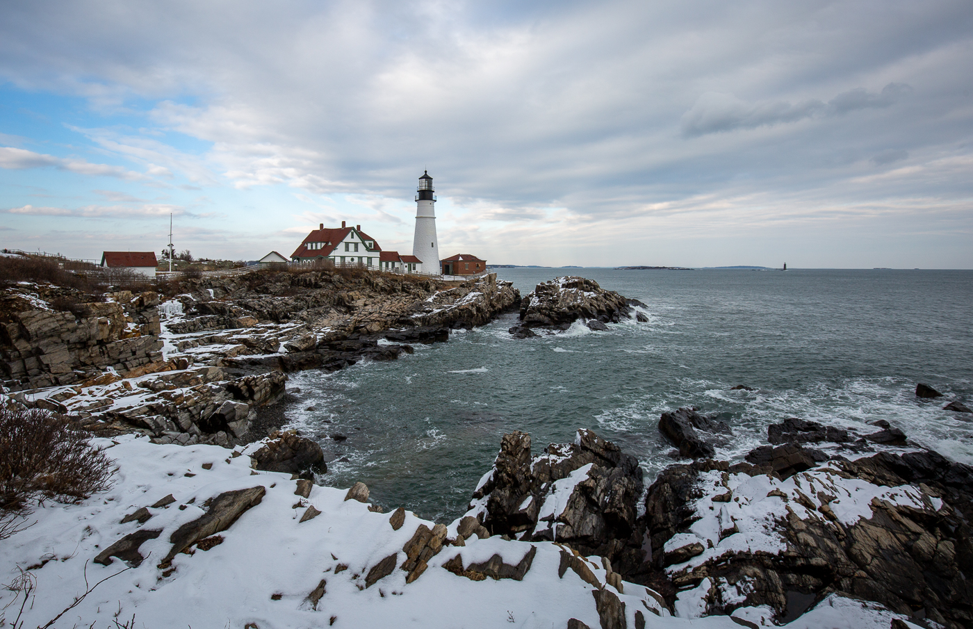 A lighthouse stands on a rocky, snow-covered coastline overlooking the ocean in Portland Maine.