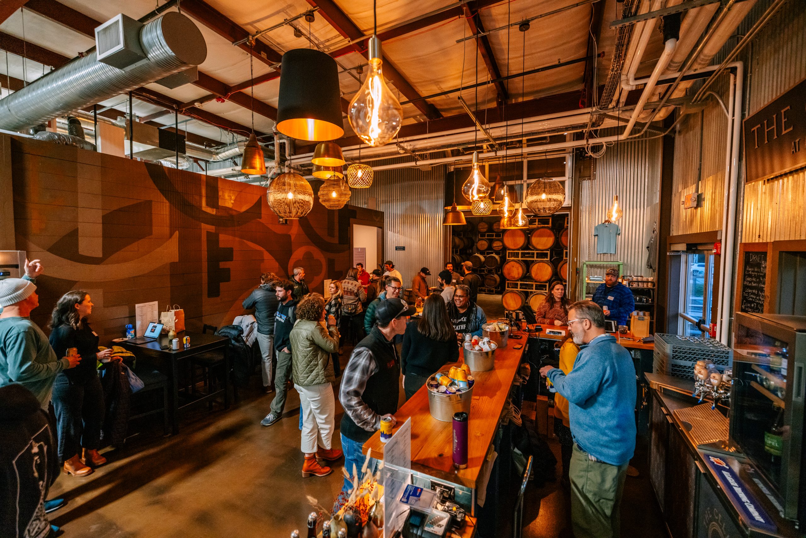 People socializing inside a rustic industrial-style brewery with wooden barrels and hanging pendant lights in Portland Maine.