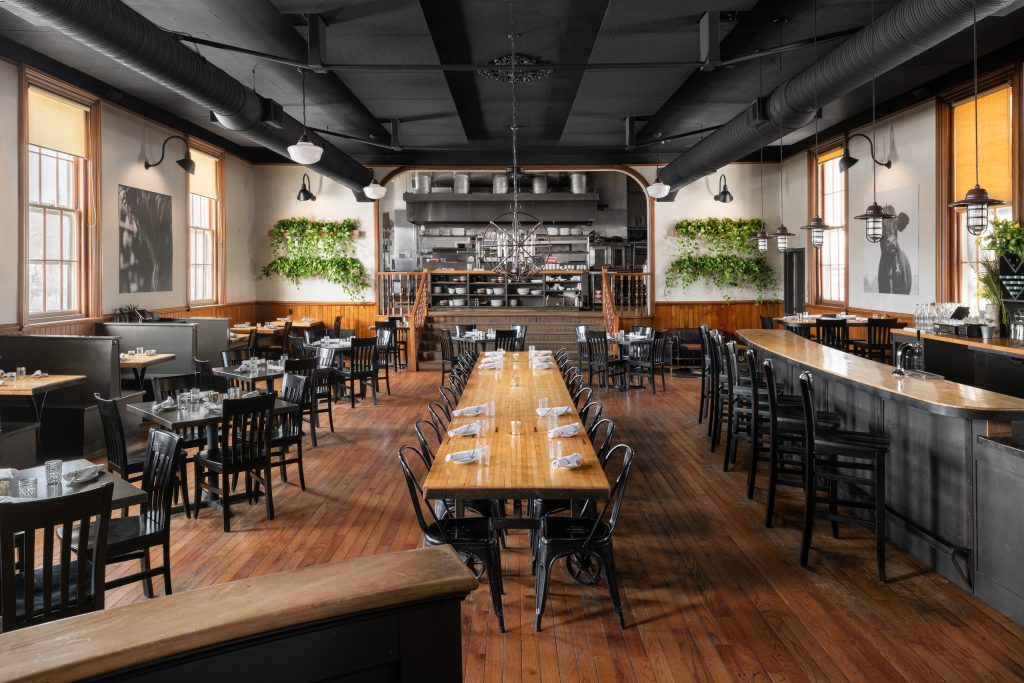 A spacious restaurant interior with wooden tables, black chairs, and an open kitchen in Portland Maine.