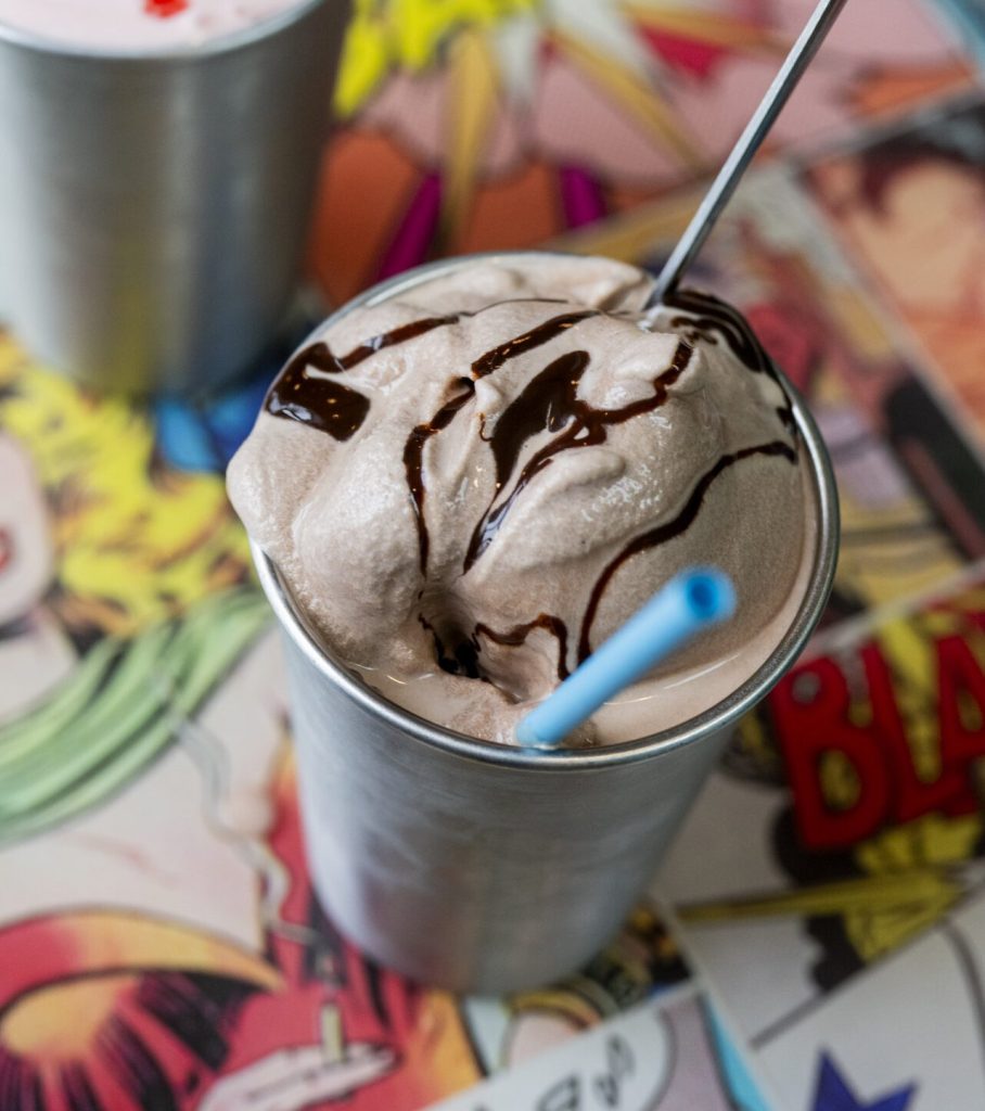 A chocolate milkshake in a metal cup with a blue straw on a comic-themed table in Portland Maine.