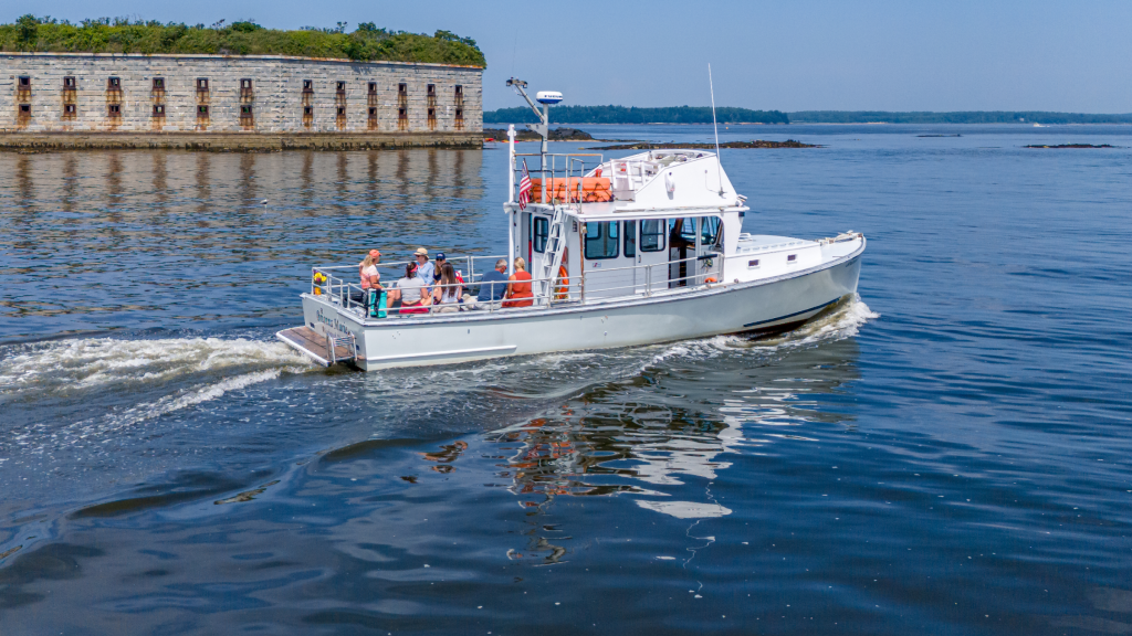 A white tour boat carries passengers past a historic stone fort on the water in Portland Maine.