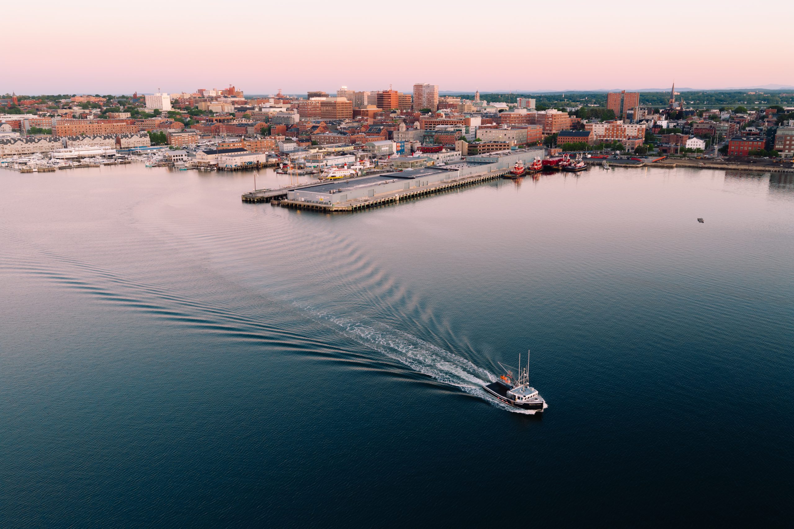 A fishing boat travels across a calm harbor toward a city waterfront at sunset in Portland Maine.