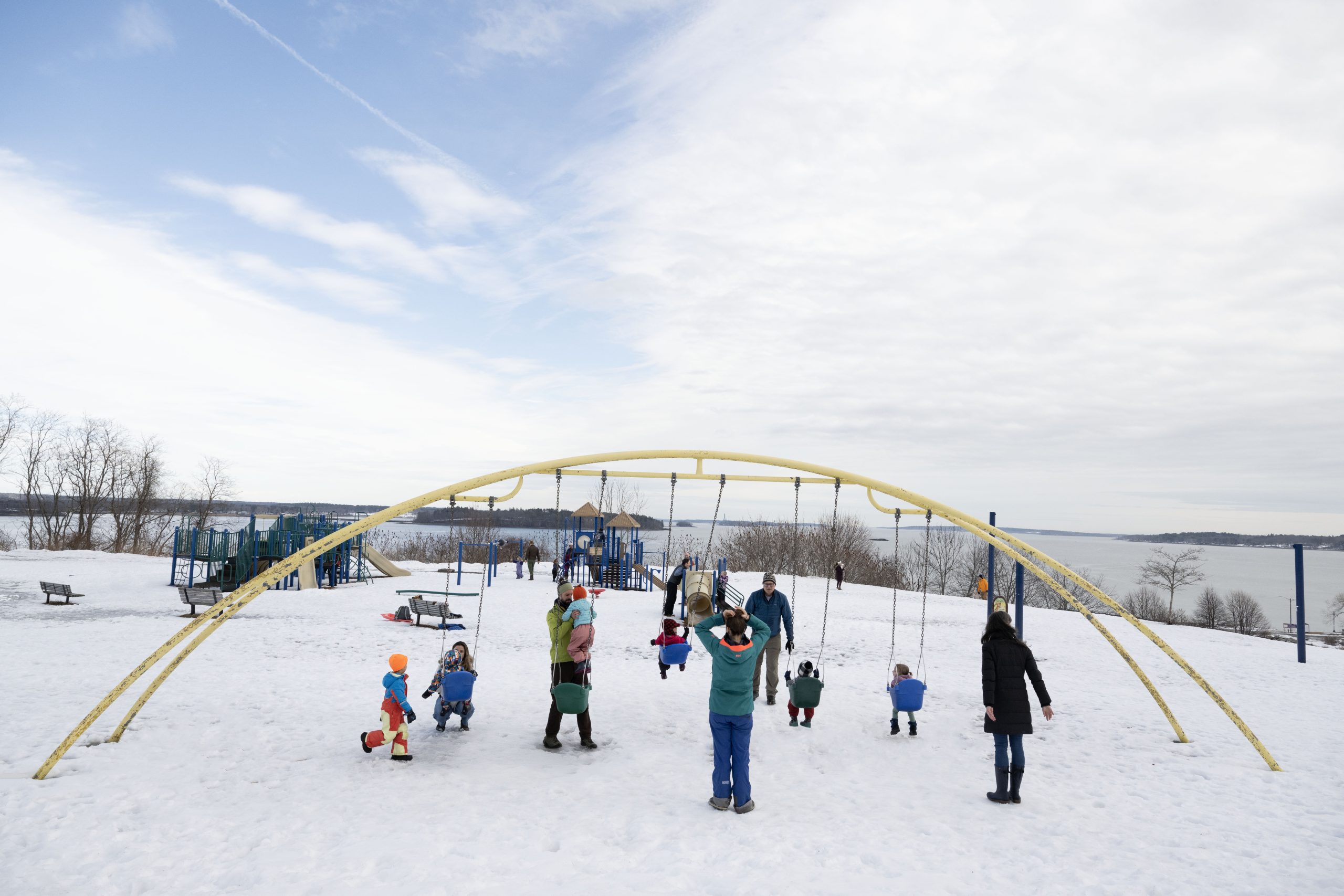 Families playing on a snowy playground with a large yellow arch swing set in Portland Maine.