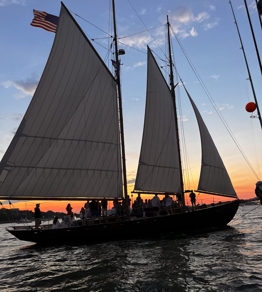 A large sailboat with passengers silhouetted against a vibrant sunset over the water in Portland Maine.