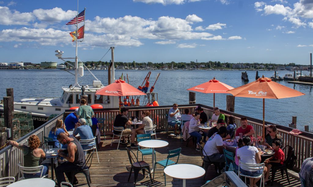 People dining on the outdoor wooden deck at Luke's Lobster overlooking a harbor with boats in Portland, Maine. Photo Credit: Serena Folding Photography