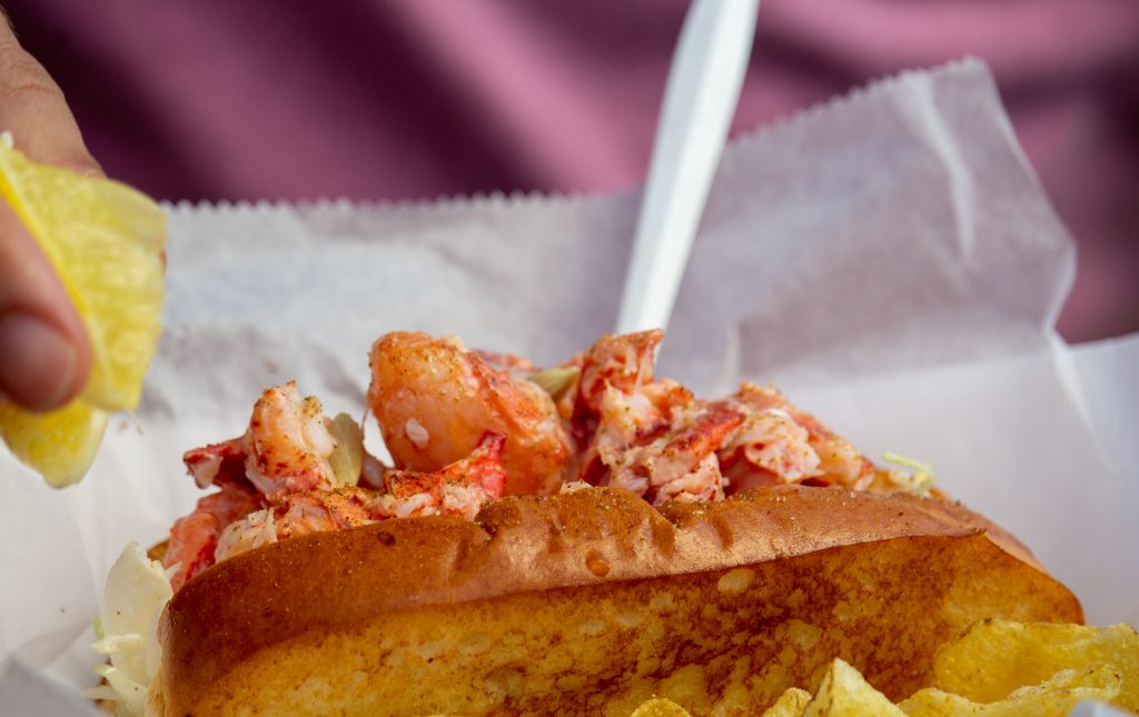 A hand squeezing a lemon wedge over a fresh lobster roll. Photo Credit: Serena Folding